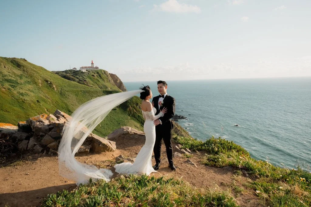 Bride and groom standing on a cliff with ocean and a lighthouse in the background, during their wedding photoshoot.