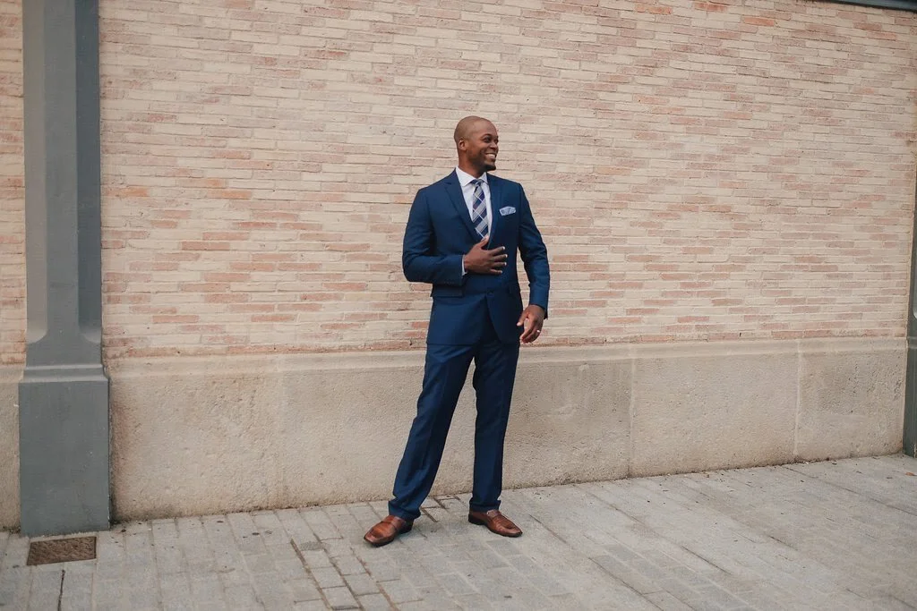 A man in a blue suit, white dress shirt, and striped tie standing on a sidewalk against a brick wall, smiling and touching his chest with his right hand.