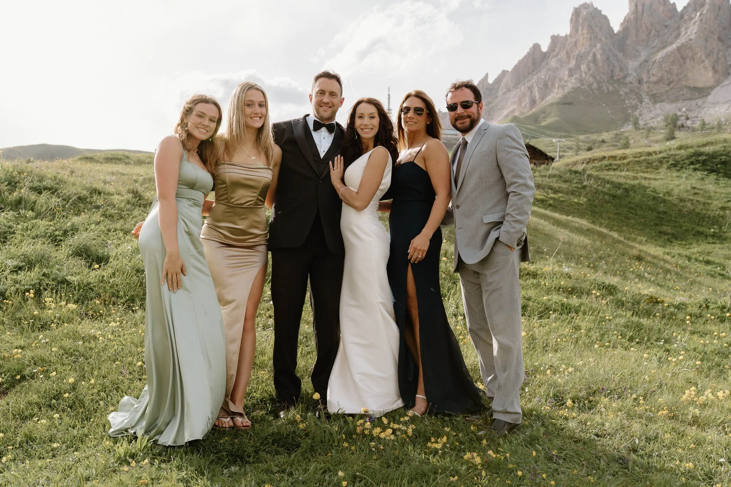 Group of six people dressed in formal attire standing on a grassy field with mountains in the background.