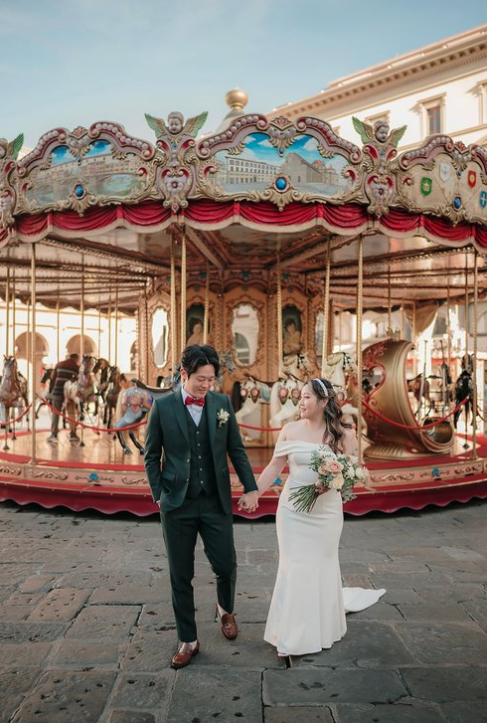 A newlywed couple holding hands and walking in front of a vintage carousel at an amusement park during daytime.