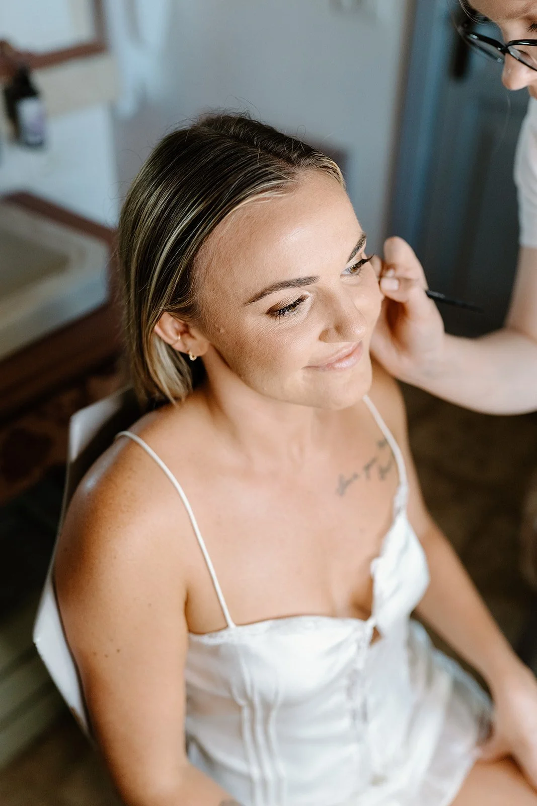 A woman with light skin and short hair is sitting while a makeup artist applies makeup to her face.