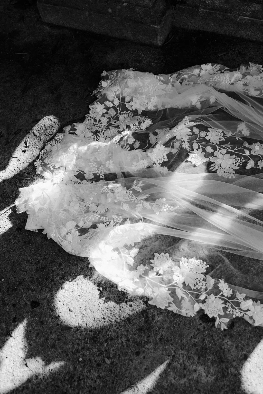 A wedding veil with floral embroidery lies on the ground near a stone wall, with sunlight casting shadows on it.