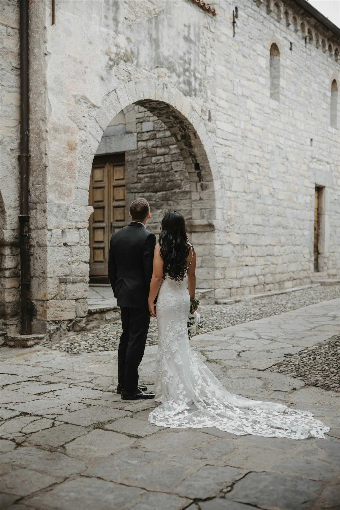 A bride and groom walking hand in hand along a cobblestone street outside a stone building with wooden doors and arched windows.