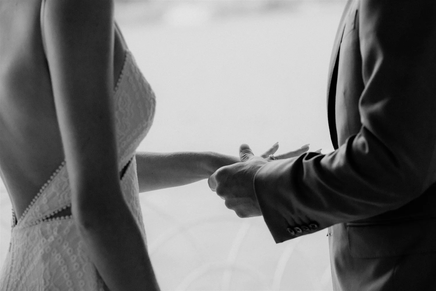 A black and white photo of a couple holding hands, with the woman in a lace dress and the man in a suit, likely during a wedding ceremony.