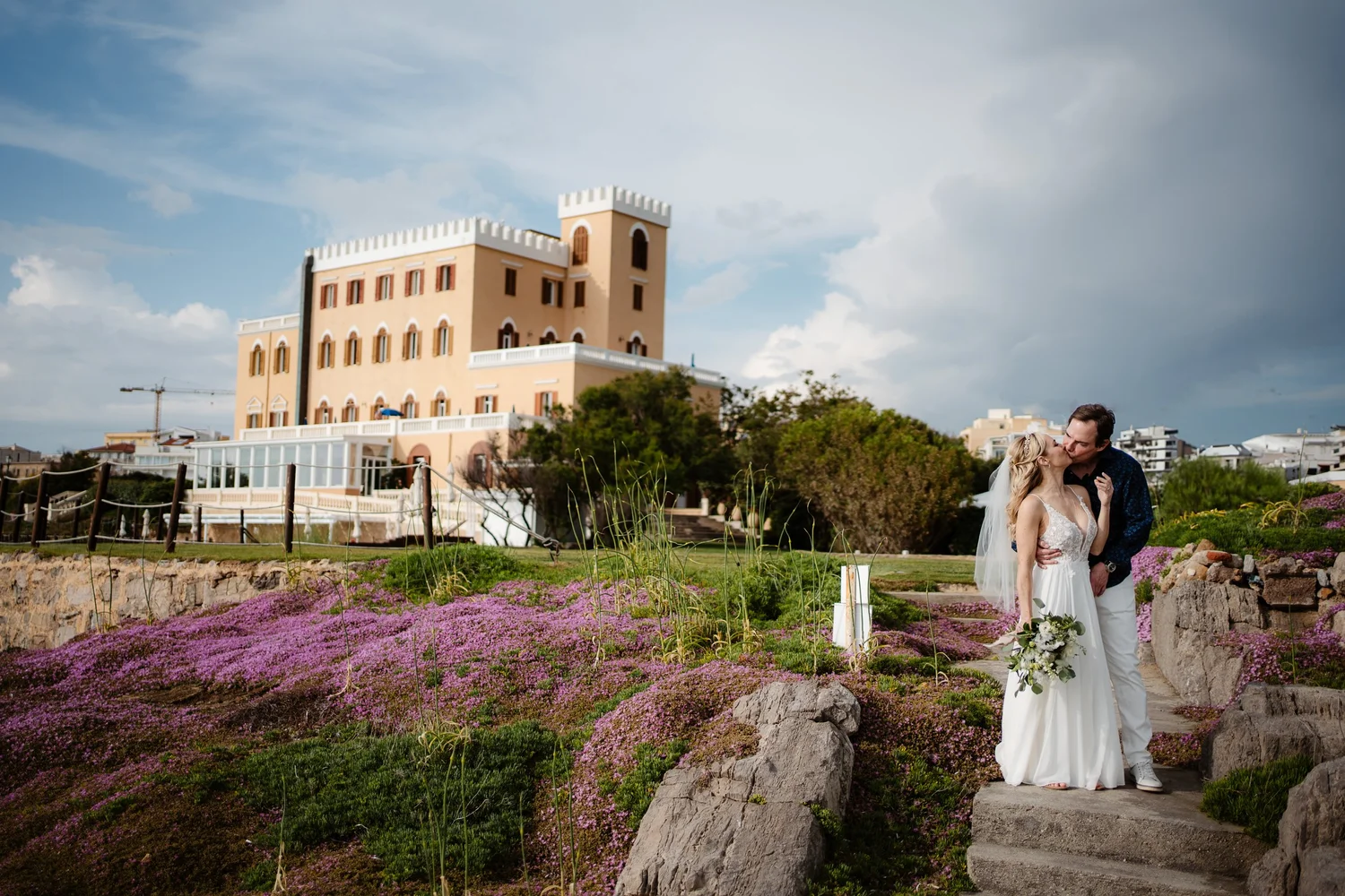 A couple in wedding attire sharing a kiss outdoors near a pink castle-like building and vibrant purple flowers.