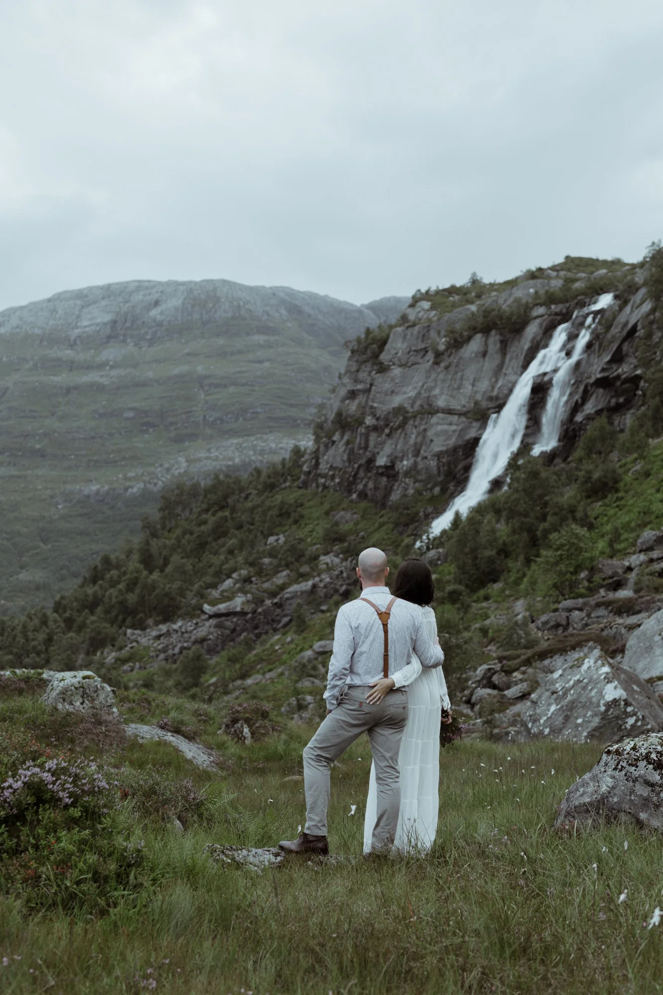A couple standing in a lush green field with rocks, facing away from the camera, with a mountain and waterfall in the background under a cloudy sky.