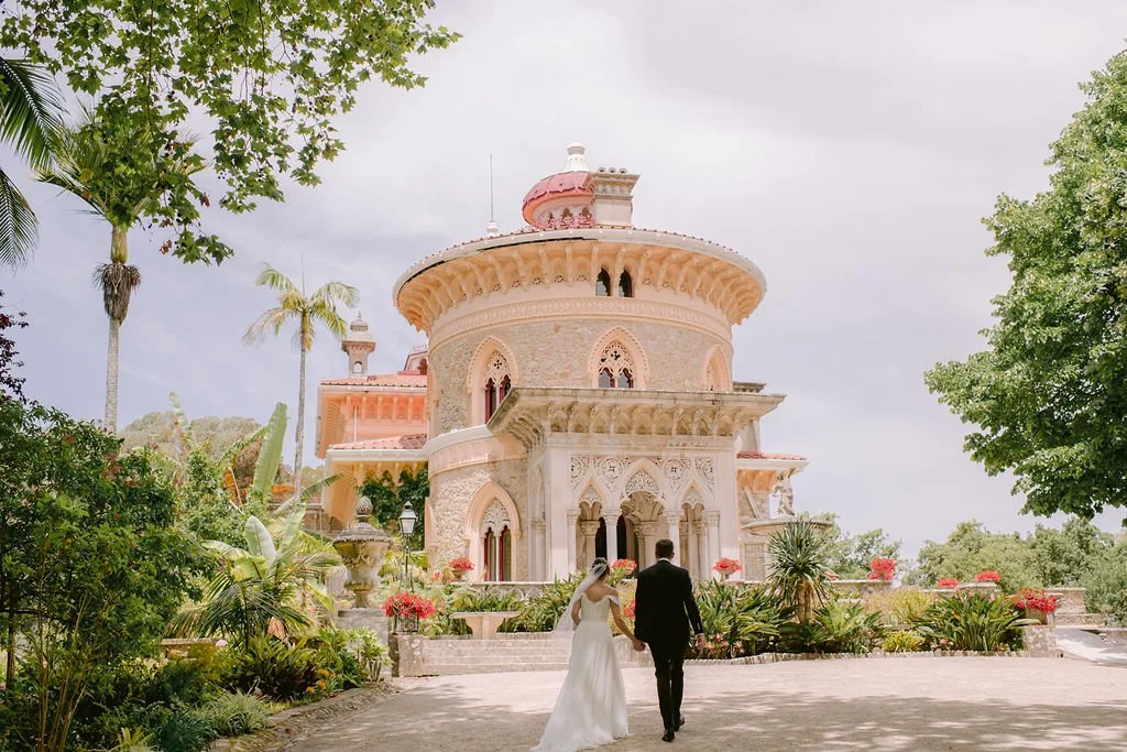 A bride and groom walking hand in hand toward a large ornate castle-style building surrounded by lush greenery and tropical plants.