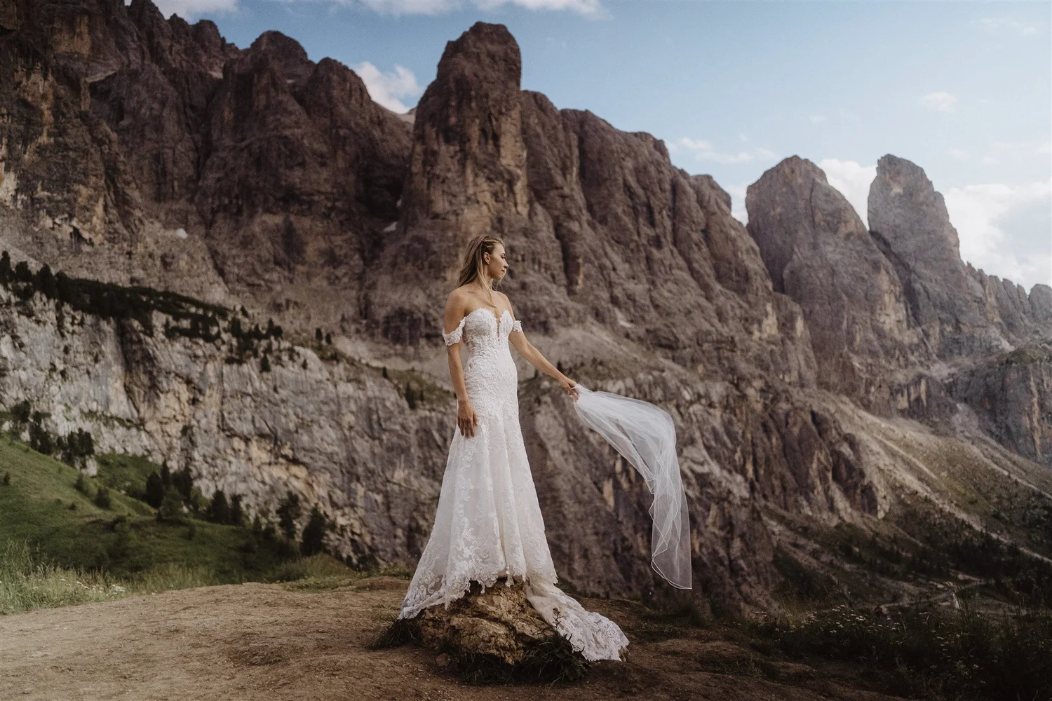 A woman in a white wedding gown standing on a rock in a mountainous landscape with rocky cliffs and a partly cloudy sky in the background.