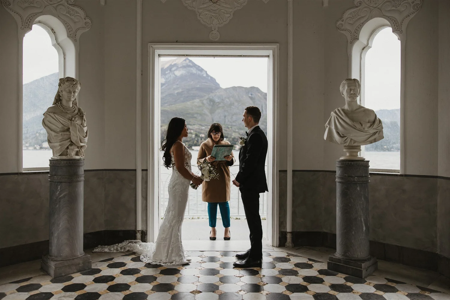 A bride and groom exchanging vows during their wedding ceremony inside a building with large windows showing mountains in the background. Two marble bust sculptures on pedestals flank the couple.