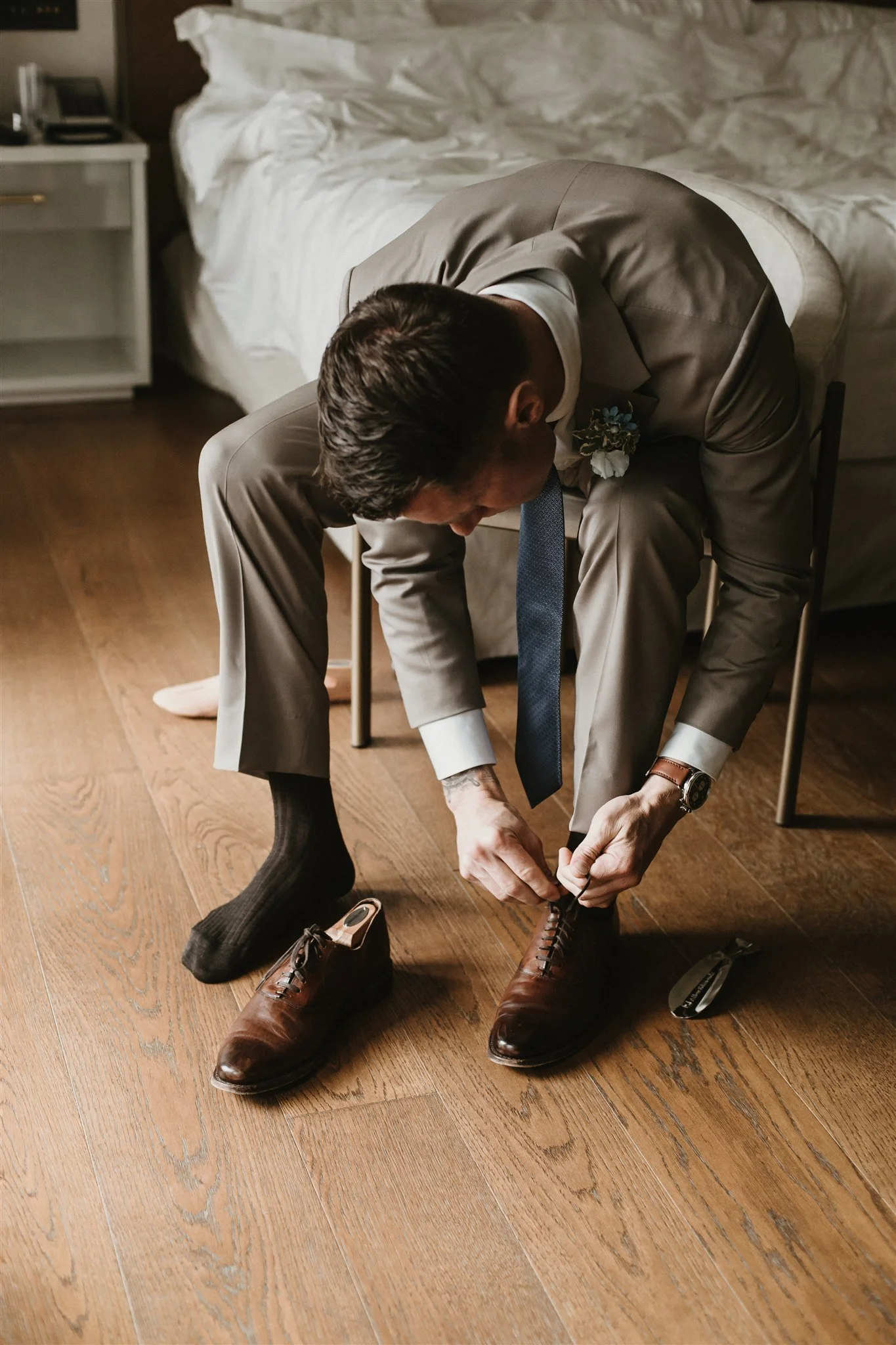 A man in a beige suit and white shirt is sitting on a chair, bending forward to tie his brown leather dress shoes. He is wearing a blue tie, a wristwatch, and has a boutonniere on his suit jacket. The room has a wooden floor, a bed with white bedding
