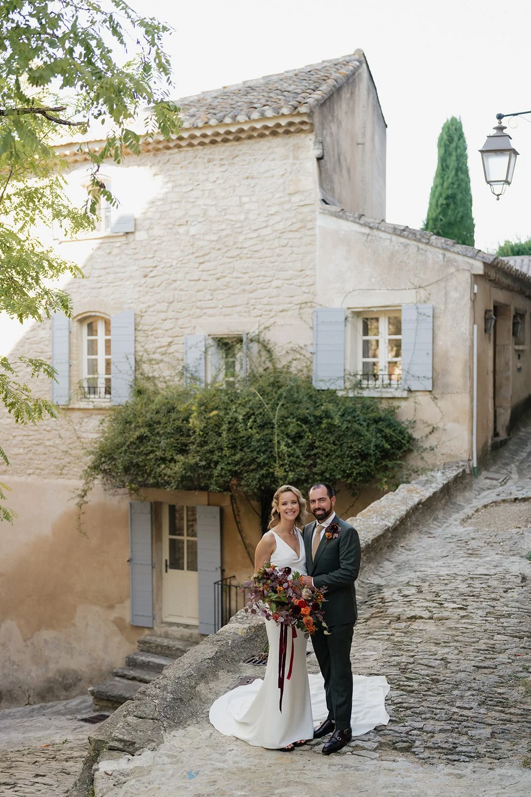 A newlywed couple standing on a cobblestone street in front of an old stone house with blue shutters, green vines, and a tree, during daytime.