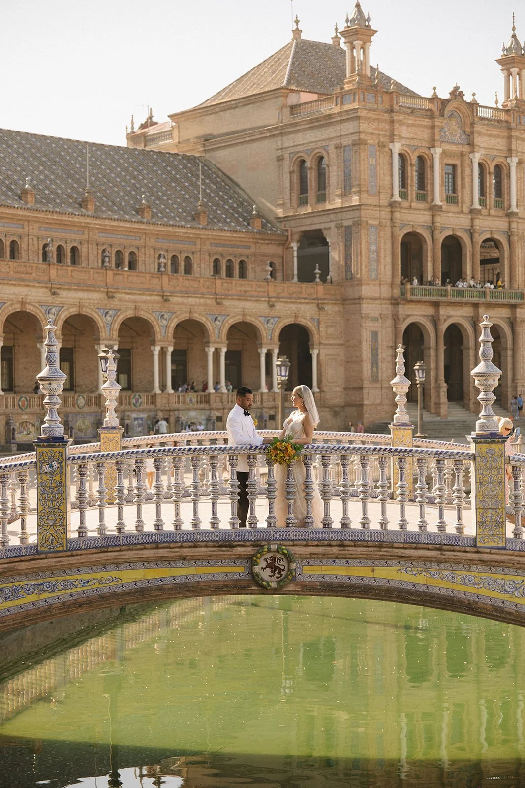 A bride and groom standing on a decorative bridge over a pond in front of a historic, ornate building during daylight with warm lighting.