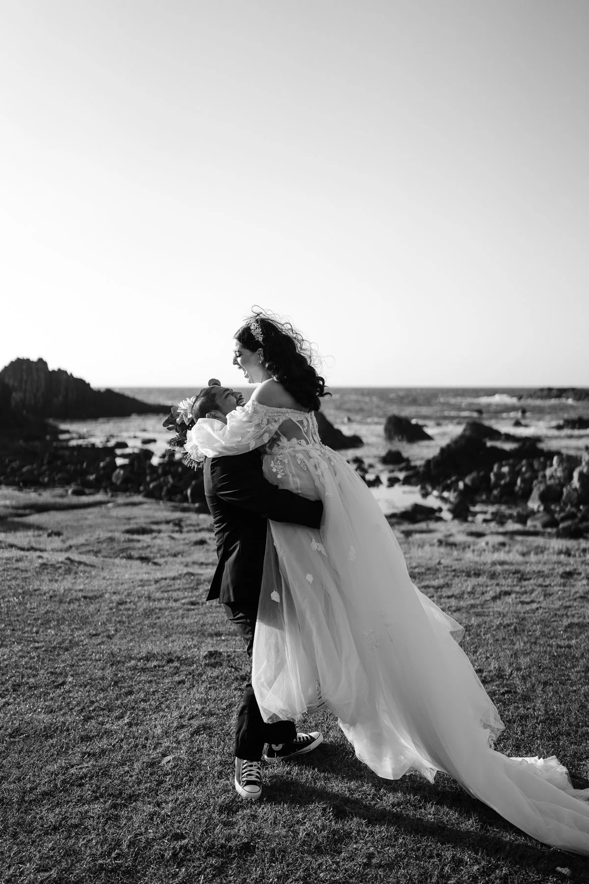 A black and white photo of a couple on the beach, with the man holding the woman in a bridal gown as they smile and look at each other.