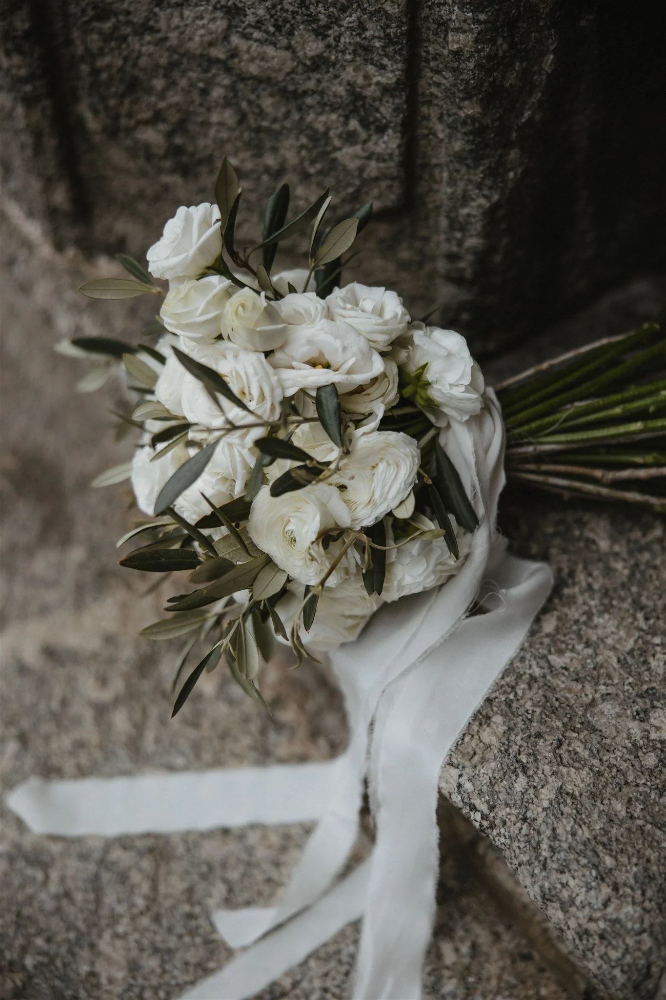 A bouquet of white flowers with green leaves tied with a ribbon, resting against a stone background.