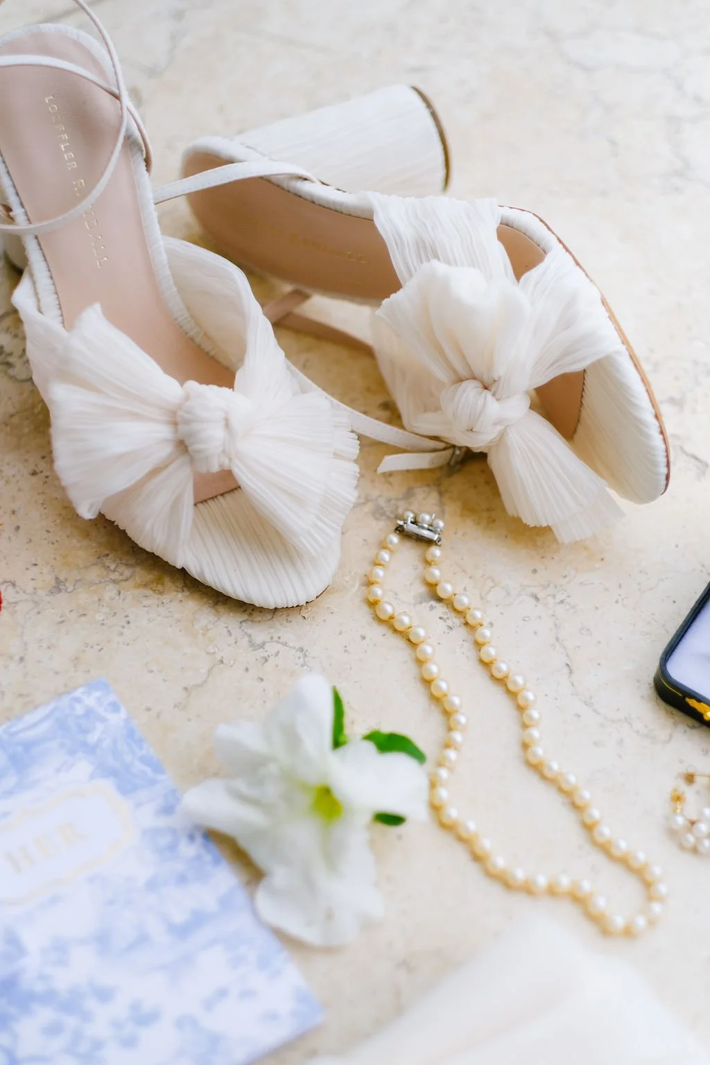 White high-heeled shoes with bow details, pearl necklace, white flower, and a cellphone on a marble surface.