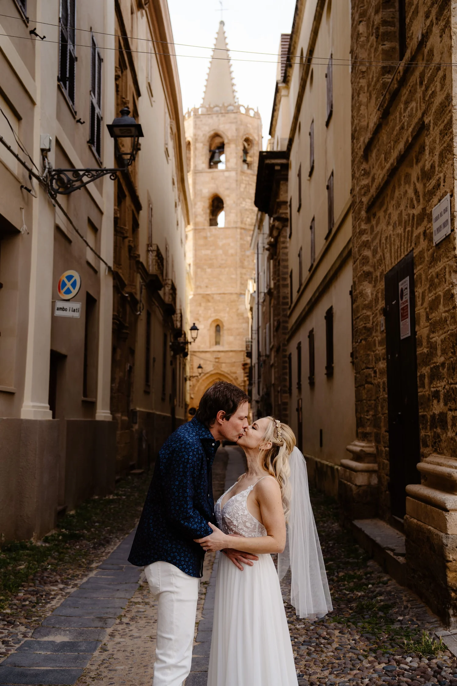 A couple in wedding attire sharing a kiss in a narrow European cobblestone street with historic buildings and a tower in the background.