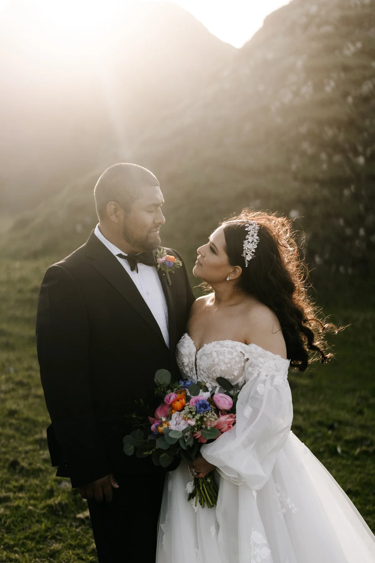 A bride and groom standing close together outdoors at sunset, gazing at each other. The groom wears a black tuxedo with a bow tie and boutonniere, and the bride wears a white strapless wedding gown with lace details and a floral headpiece. The bride