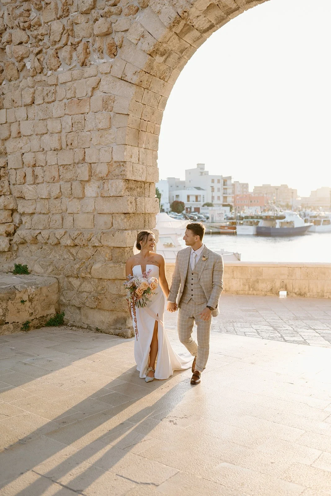 A bride and groom walking hand in hand by the waterfront at sunset, with boats and buildings in the background, under an ancient stone arch.
