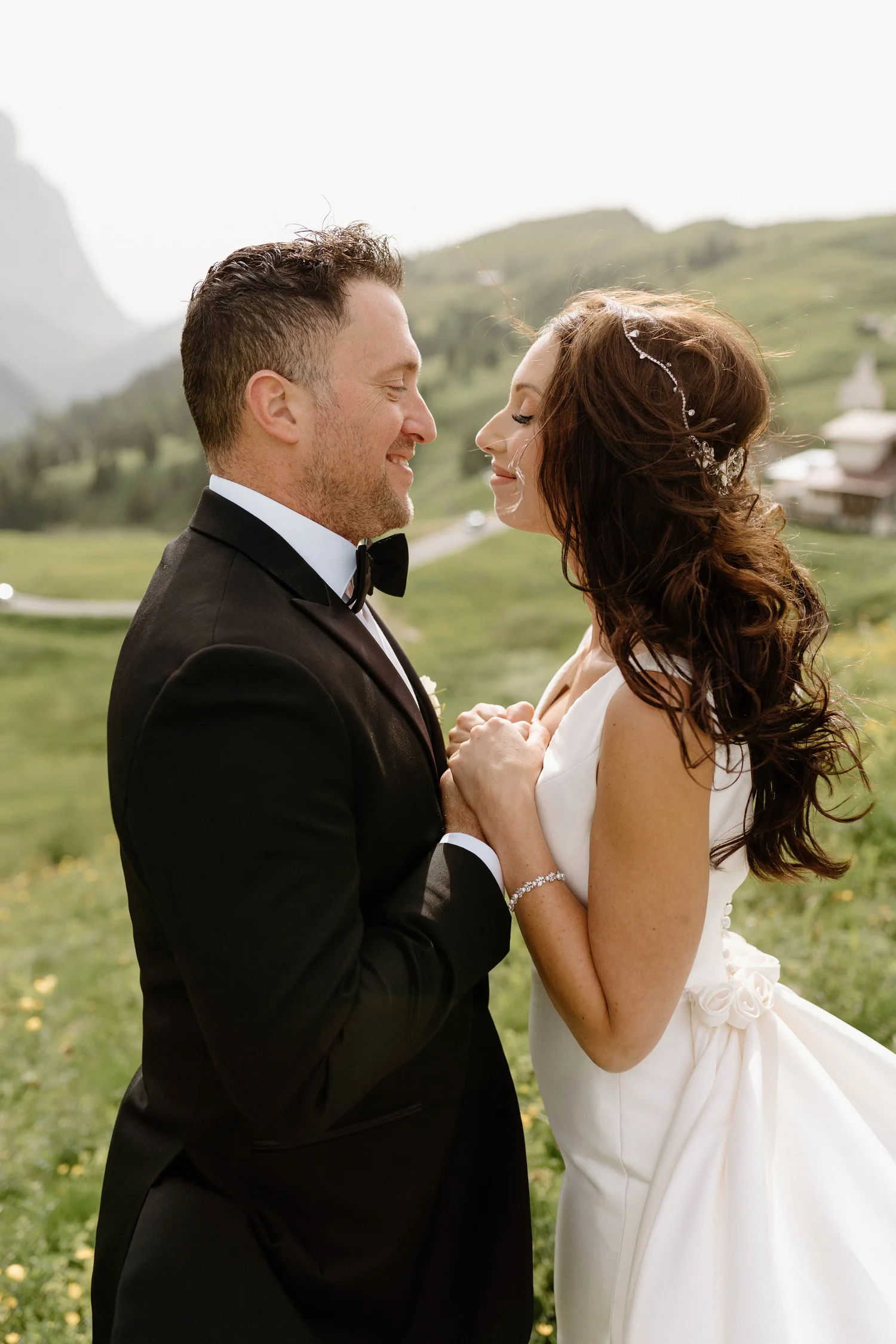 A bride and groom on their wedding day, holding hands and smiling in an outdoor green setting with a mountain and buildings in the background.
