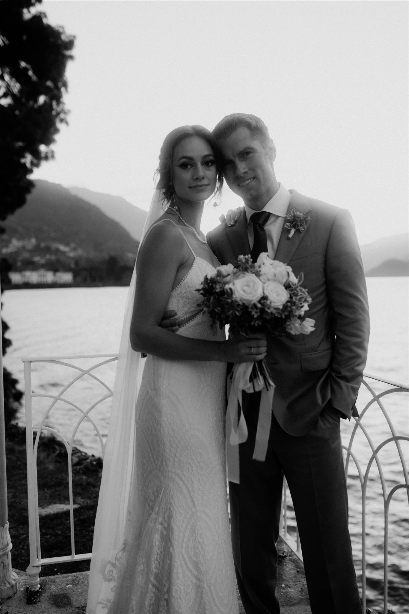 A bride and groom standing close together outdoors near water, with a hill and trees in the background. The bride is holding a bouquet and wearing a lace wedding dress, while the groom is in a suit with a boutonniere, smiling for the photo.