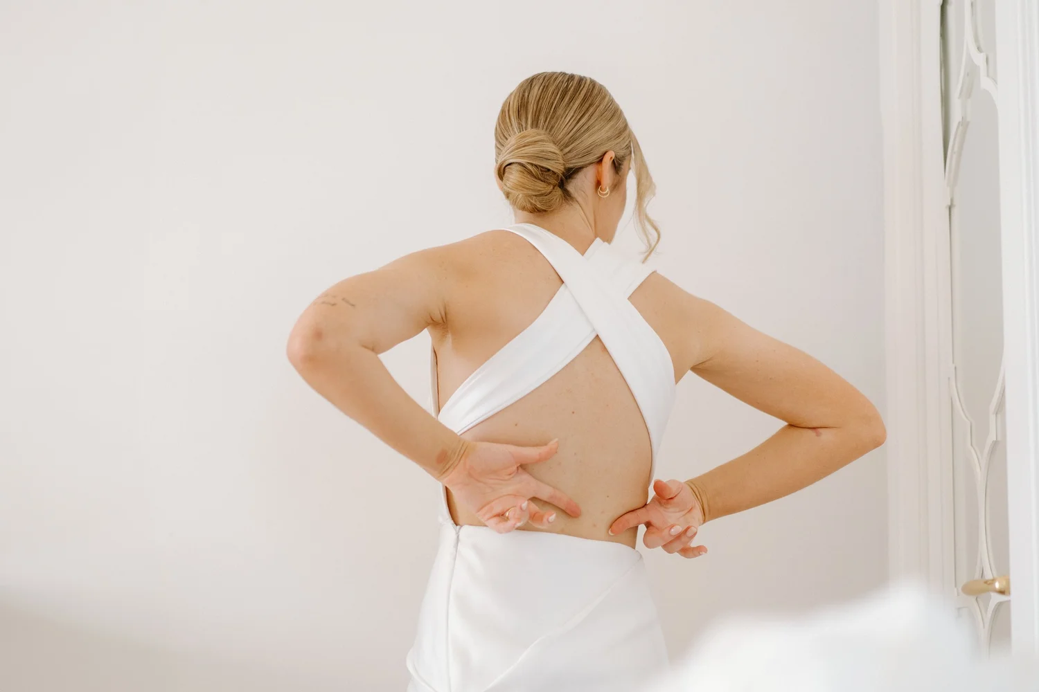 A woman with blonde hair in an elegant updo, wearing a white sleeveless dress, stands with her back to the camera in front of a mirror with decorative framing. She is touching her side with one hand and resting her other hand on her hip, looking into