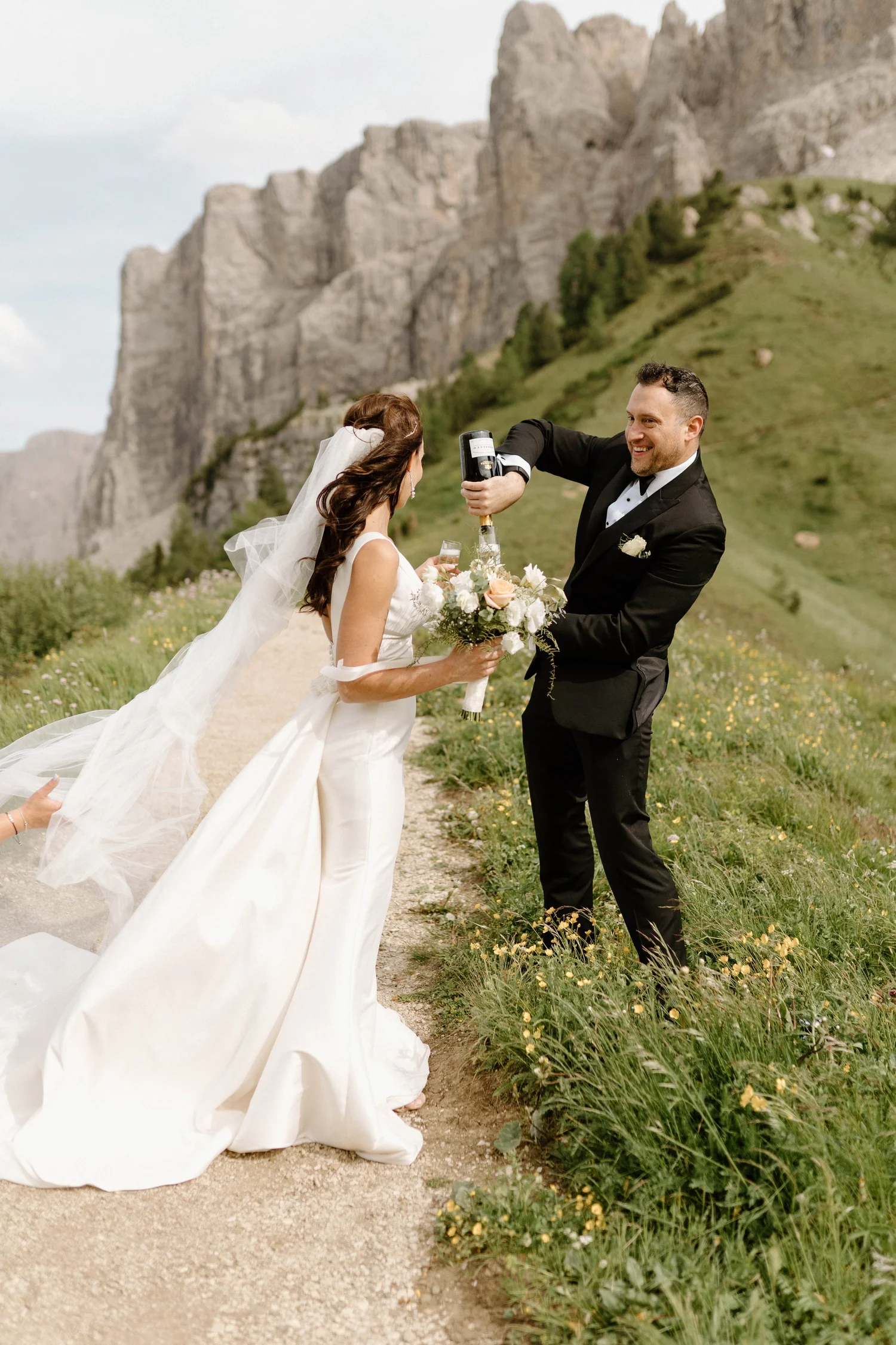 A bride and groom celebrating outdoors on a wedding day. The groom is pouring champagne into the bride's glass while they smile at each other. The bride is holding a bouquet of flowers, and the background features green hills and rocky cliffs.