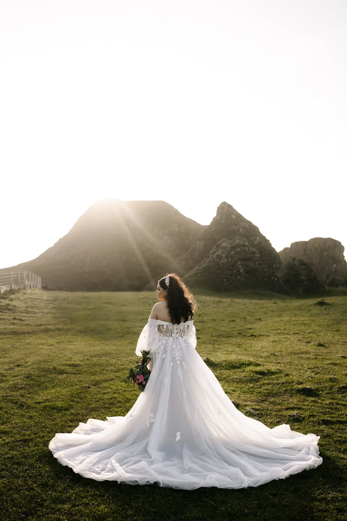A woman in a white wedding dress holding a bouquet of flowers stands on a grassy field with mountains in the background during sunset.