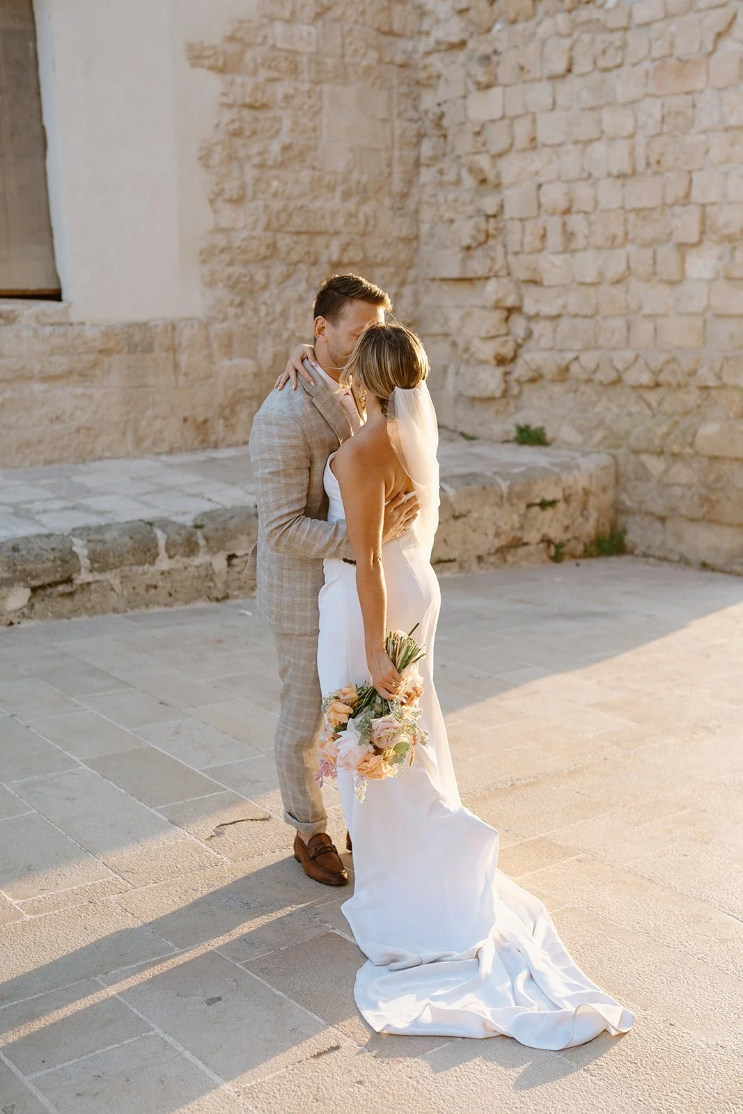 A bride and groom embrace outdoors during sunset, with the bride holding a bouquet of flowers, against an old stone wall.