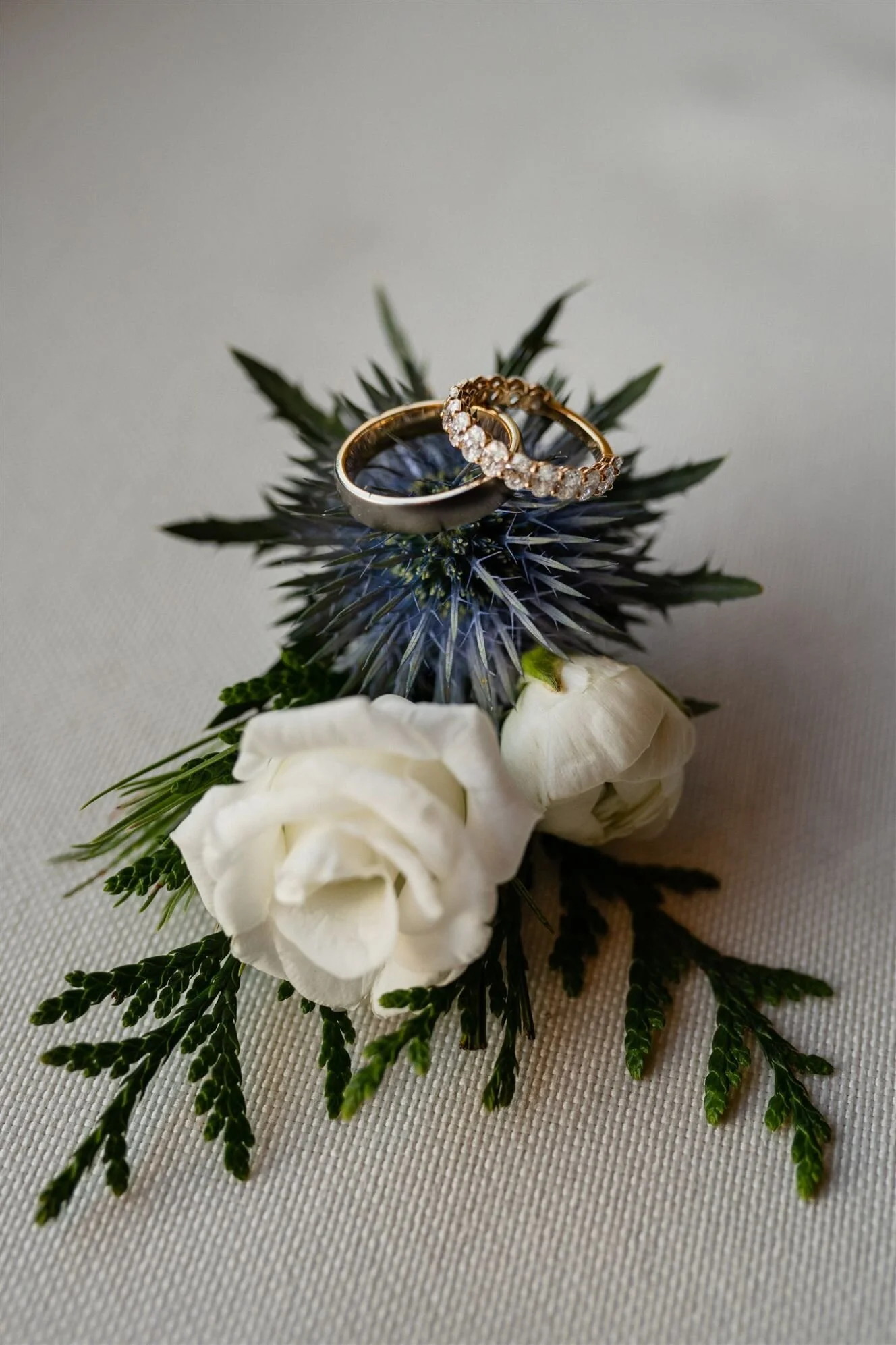 Wedding rings, one band with diamonds and a plain band, resting on a thistle flower with white roses and greenery.