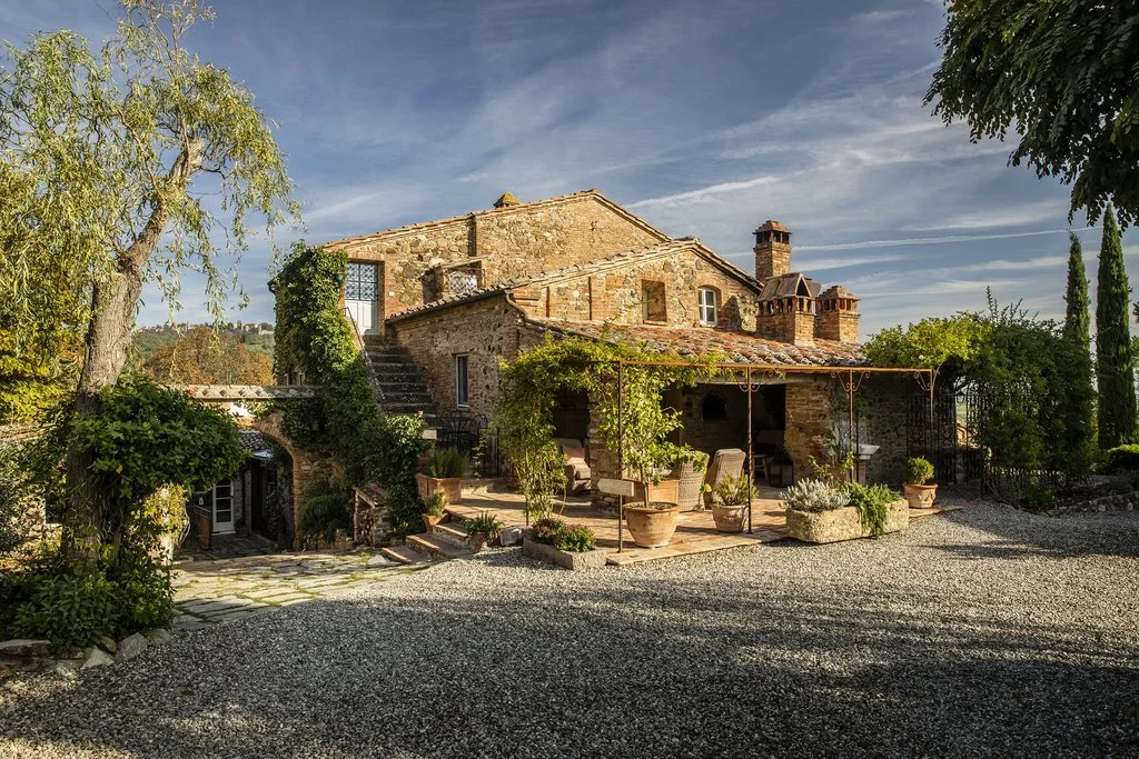 A rustic brick house with a terracotta tile roof, surrounded by greenery and potted plants, under a blue sky with some clouds.