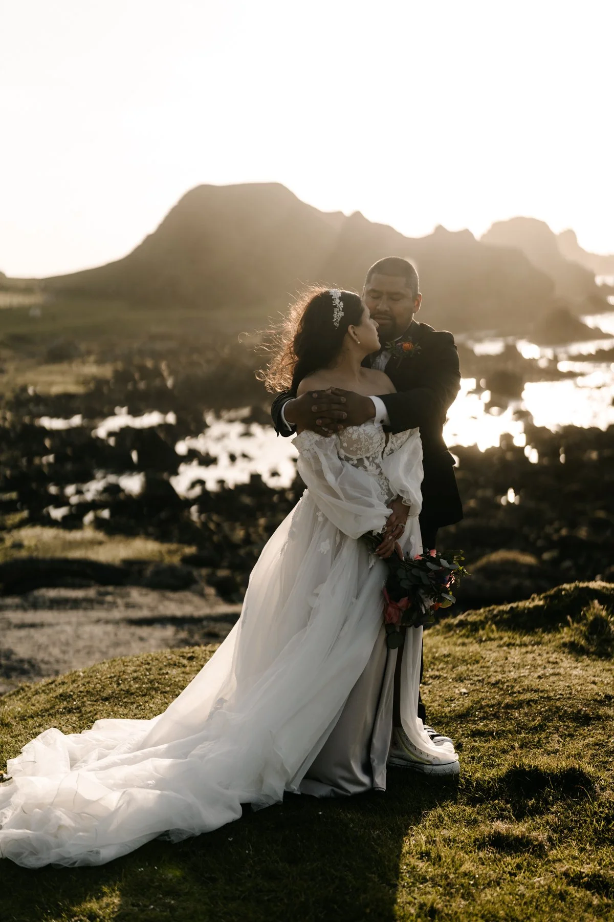 A bride and groom embracing outdoors during sunset with rocky hills and water in the background.