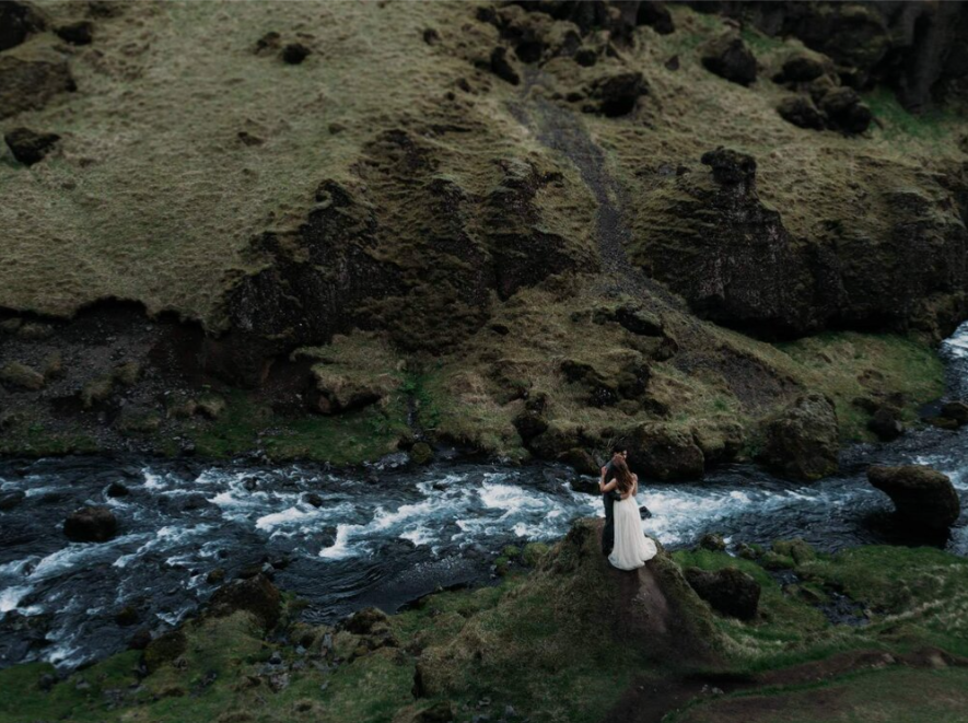 Couple standing on rocks by a rushing river in a green, hilly landscape.