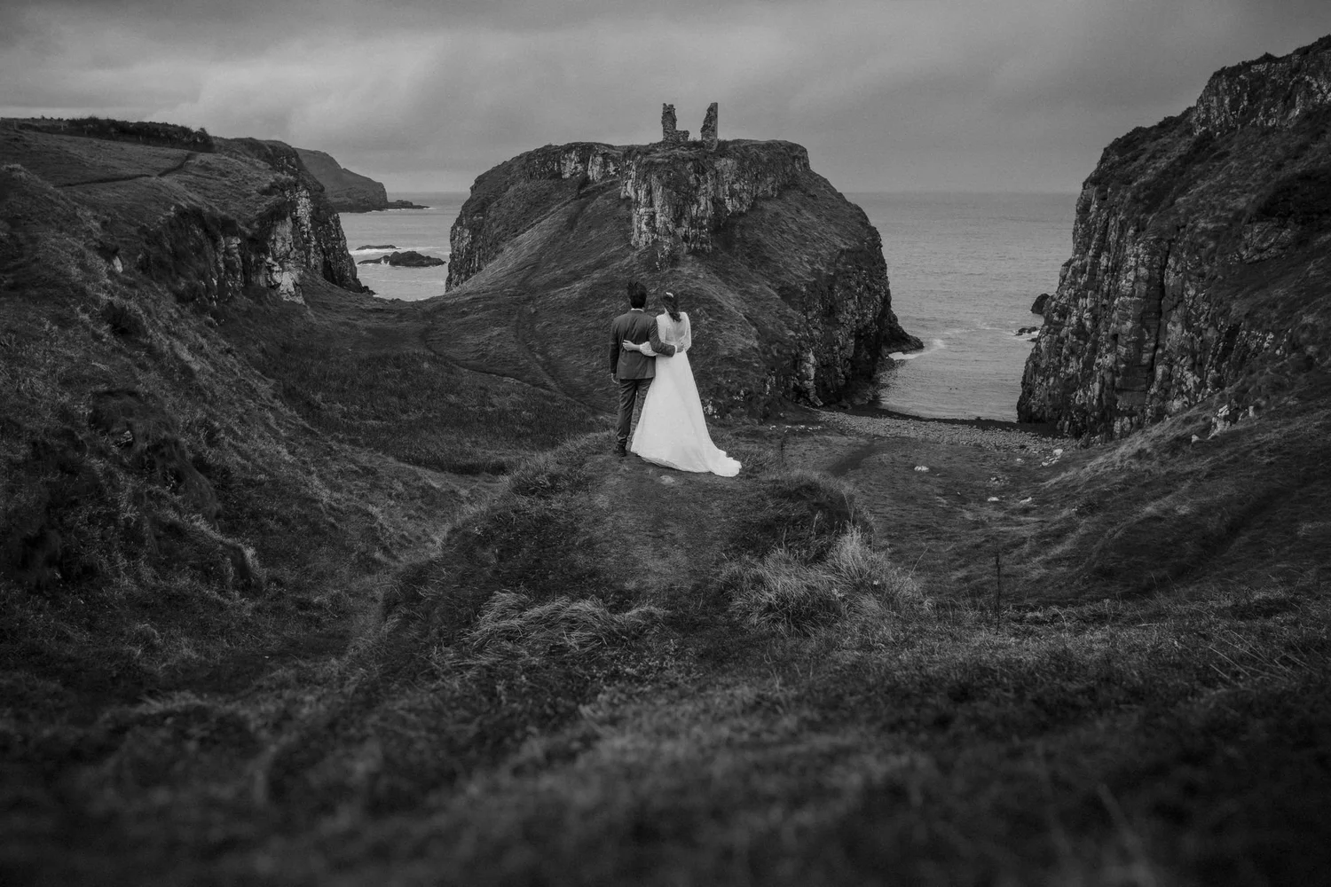 A black and white photograph of a couple, dressed in wedding attire, standing on a grassy hill near the coast, with large rocky cliffs and a castle ruin in the background.