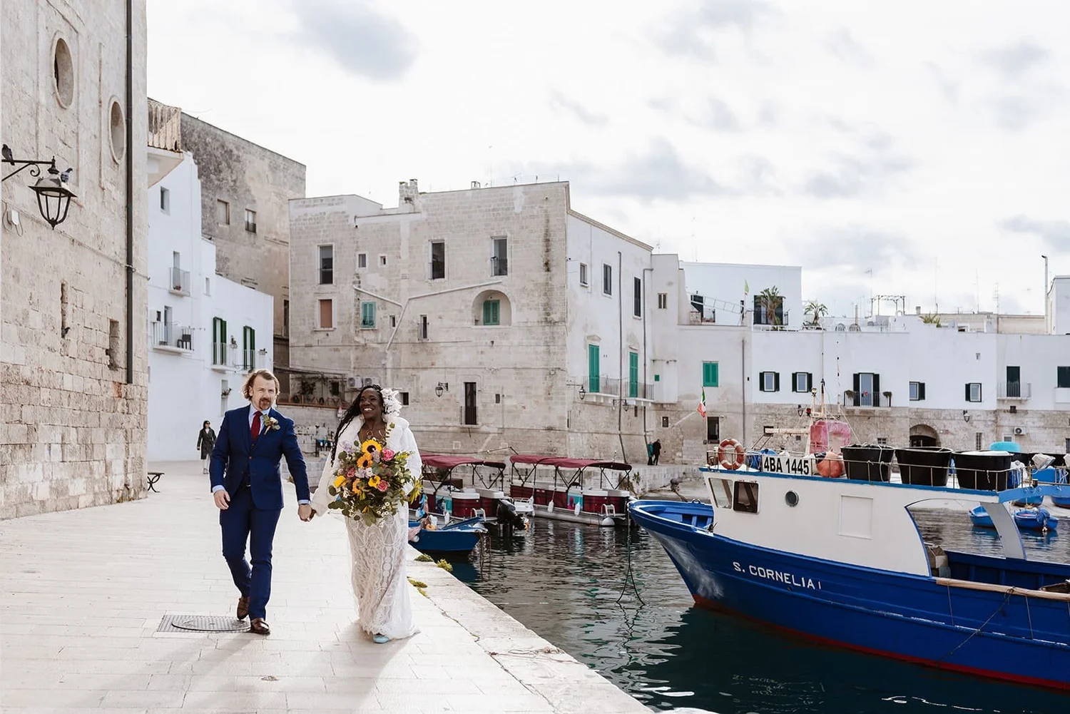 A newlywed couple walking by a harbor with boats, historic white buildings, and cloudy sky in the background.