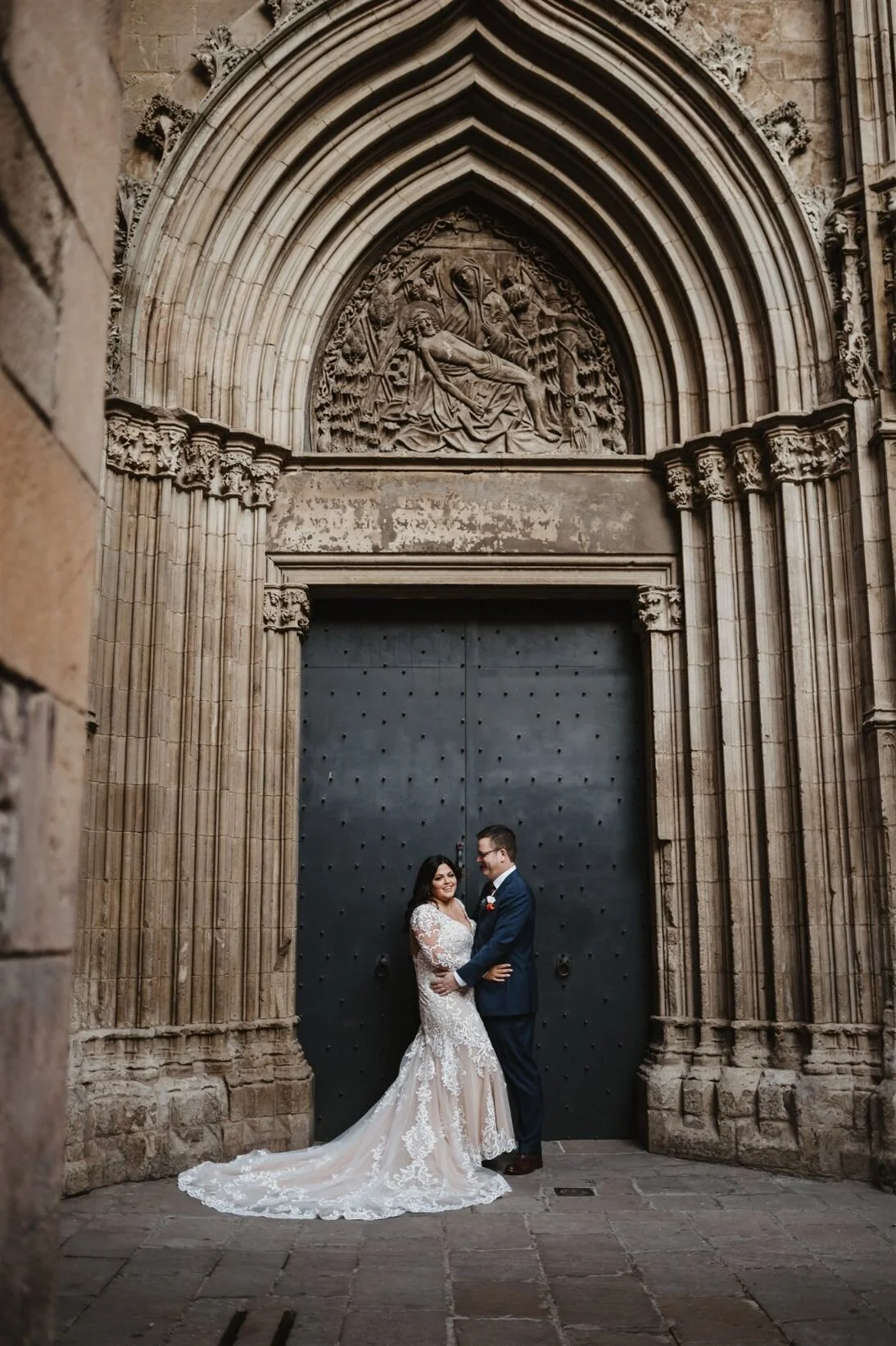 A newlywed couple in wedding attire standing in front of a large, ornate church door with intricate stone carving above it, smiling at each other.
