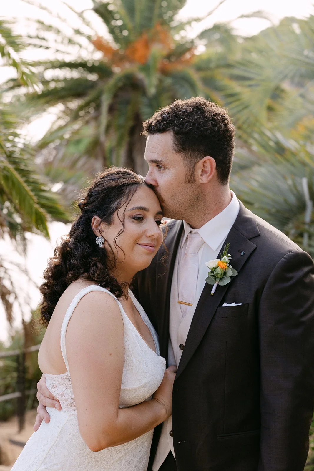 A newlywed couple shares an intimate moment outdoors, with the groom kissing the bride’s forehead in front of palm trees.