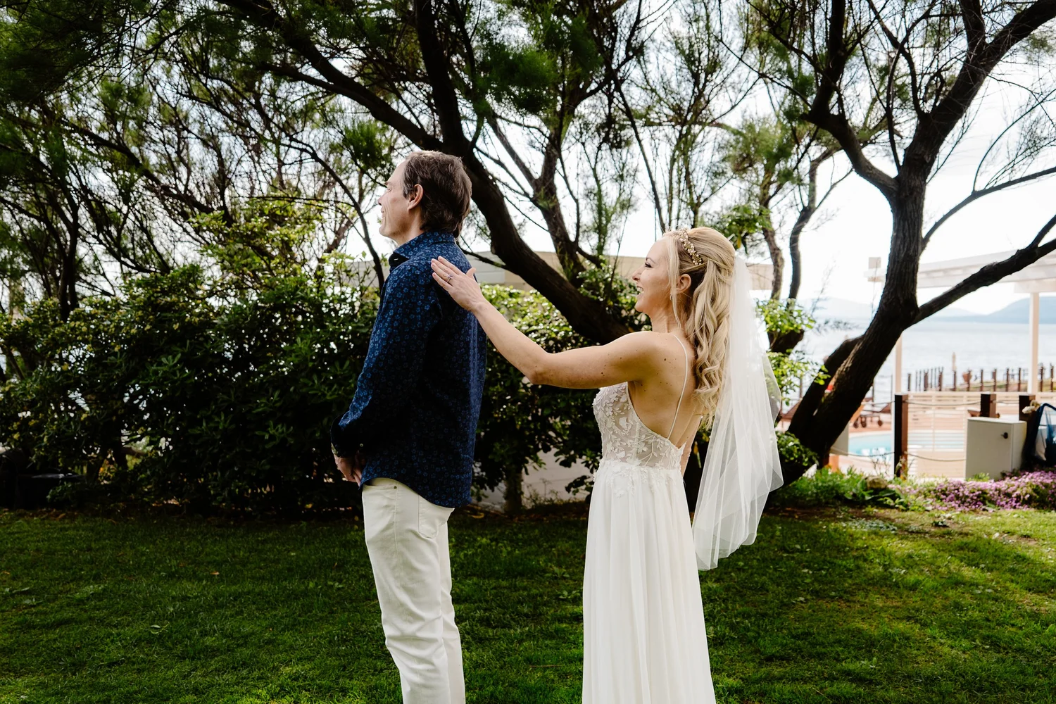 A bride and groom on their wedding day outdoors, with the bride smiling and touching the groom's shoulder, behind a large tree and greenery, with water and a pier in the background.