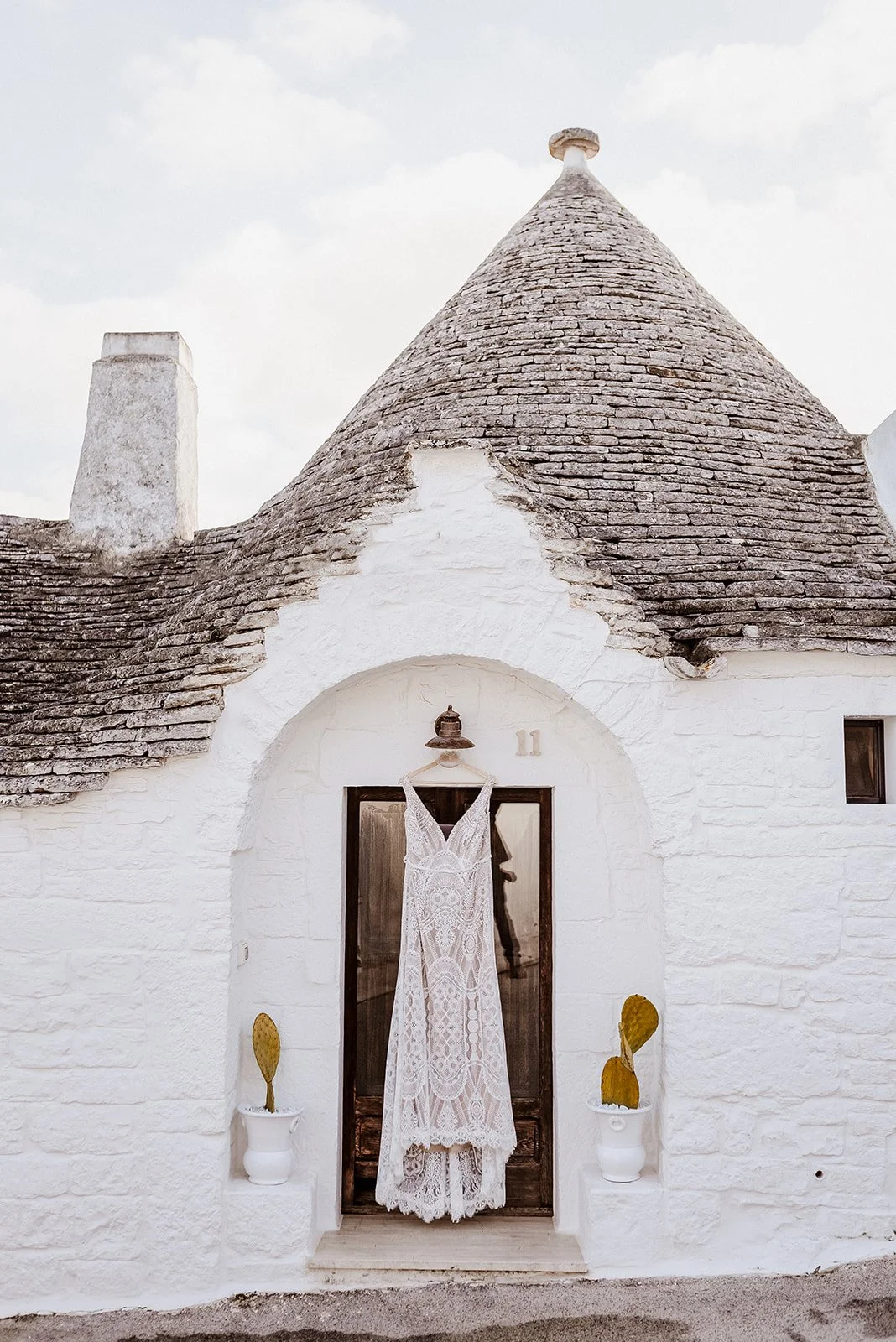 White house with a conical roof and a wooden door, a white lace wedding dress hanging on the door, two yellow cactus plants in white pots on either side, and a house number 11 above the door.