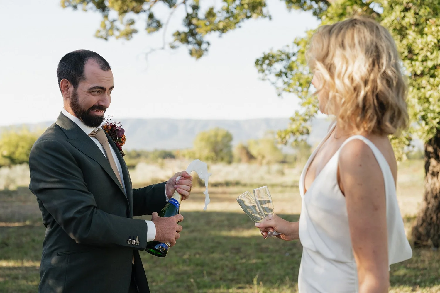 A man in a suit opening a bottle of champagne with foam coming out, celebrating with a woman in a white dress holding two glasses of champagne, outdoors under trees with a scenic landscape in the background.