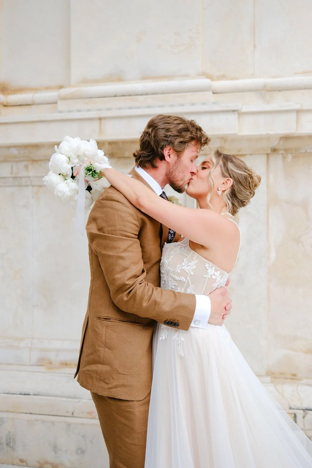 A bride and groom share a kiss on their wedding day, with the bride in a white dress holding a bouquet of white flowers and the groom in a brown suit, standing in front of a stone wall.