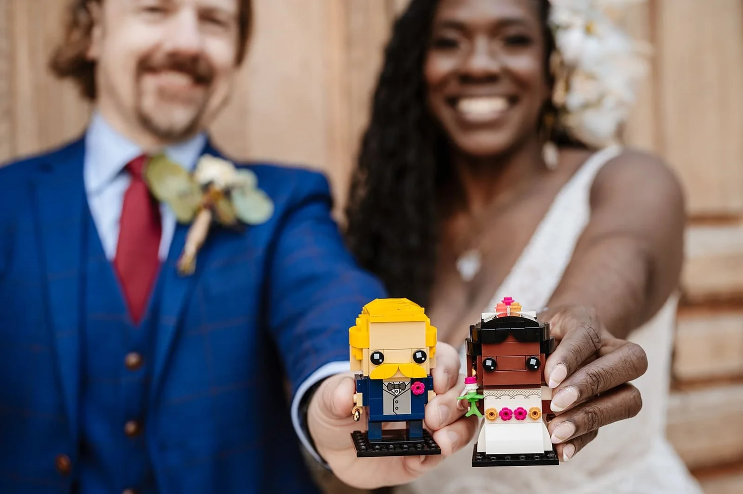 A smiling couple holding LEGO figurines of a groom and a bride. The man is wearing a blue suit with a boutonniere, and the woman is wearing a wedding dress with a pearl necklace and flower earrings.