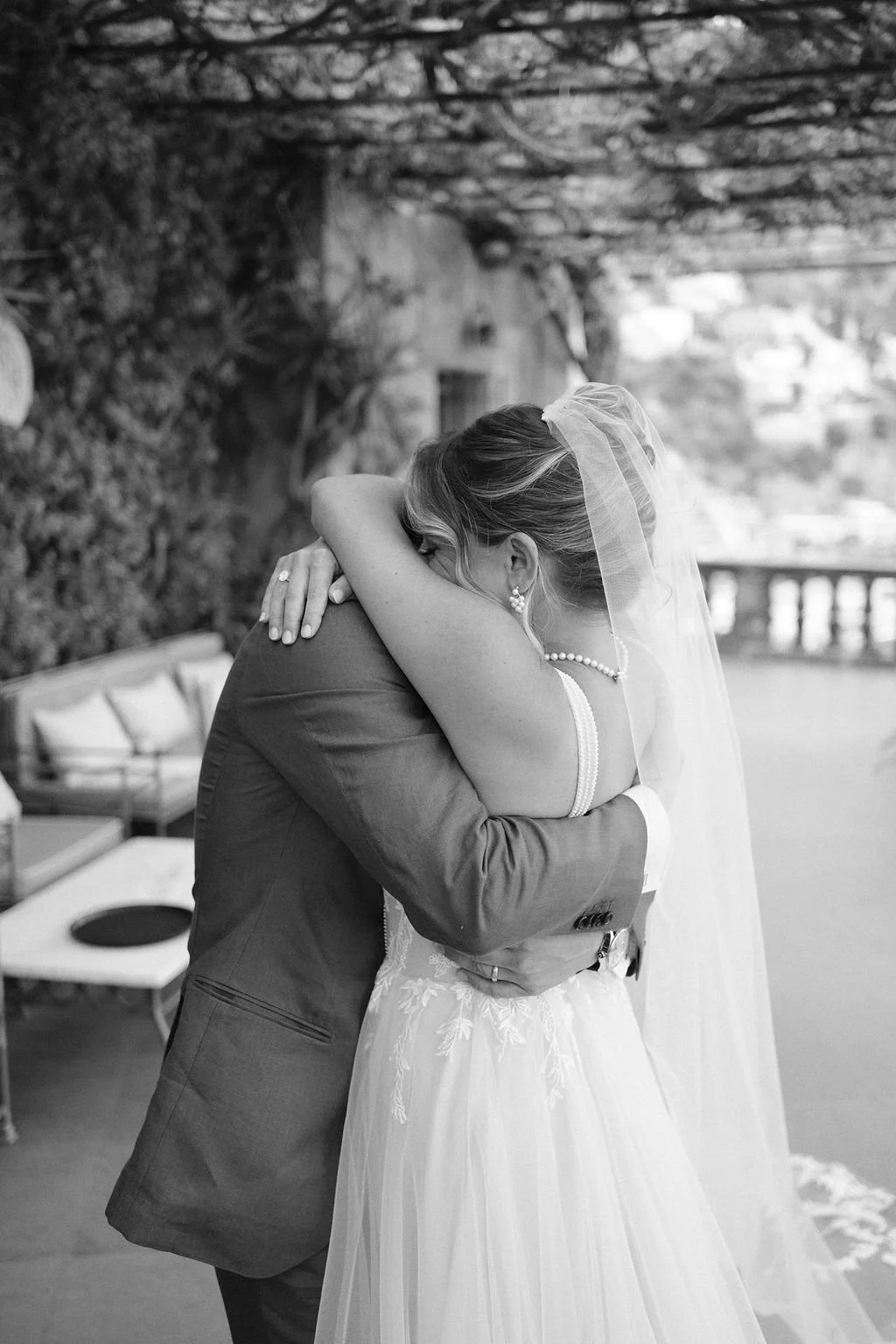 A bride and groom embrace during their wedding, with the bride wearing a wedding gown and veil, and the groom in a suit, on an outdoor terrace with trees and cityscape in the background.