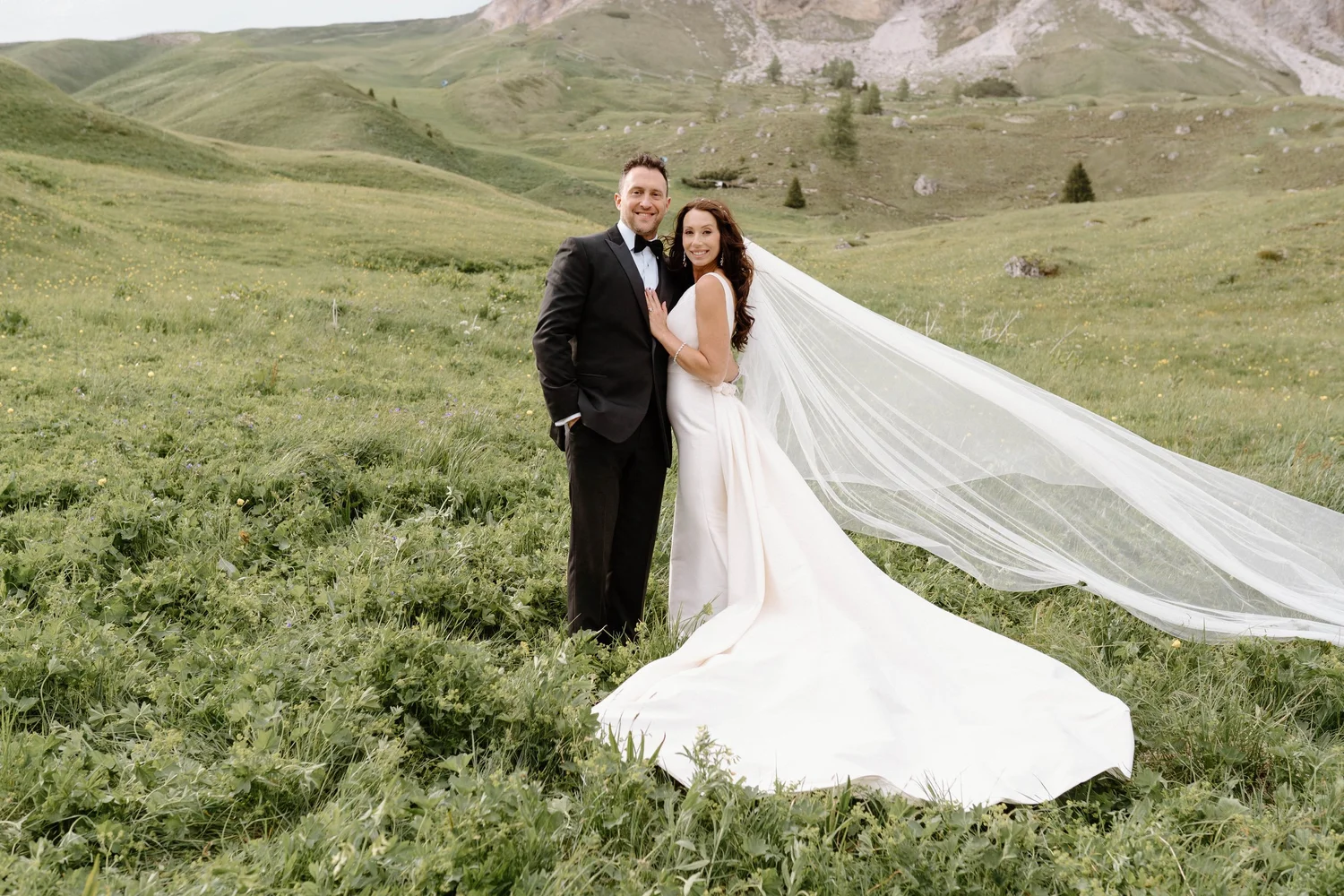 A newlywed couple in wedding attire standing on a green meadow with hills and mountains in the background, smiling at the camera.