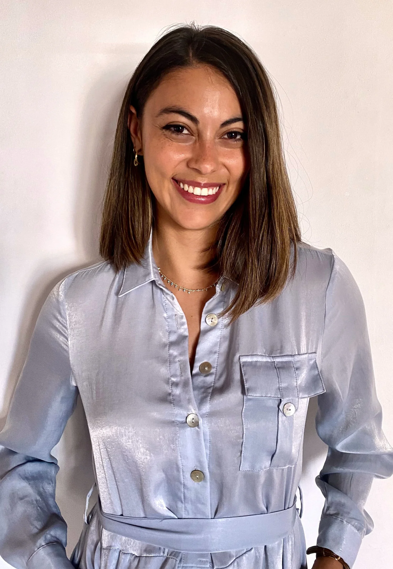 A woman with shoulder-length brown hair, wearing a shiny silver button-up shirt, silver skirt, earrings, a necklace, and a watch, smiling and standing against a plain, light-colored wall.