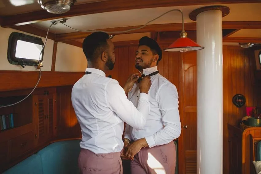 Two men in white shirts and pink pants adjusting each other's ties inside a wooden-paneled room, with a red lampshade and a white column in the background.