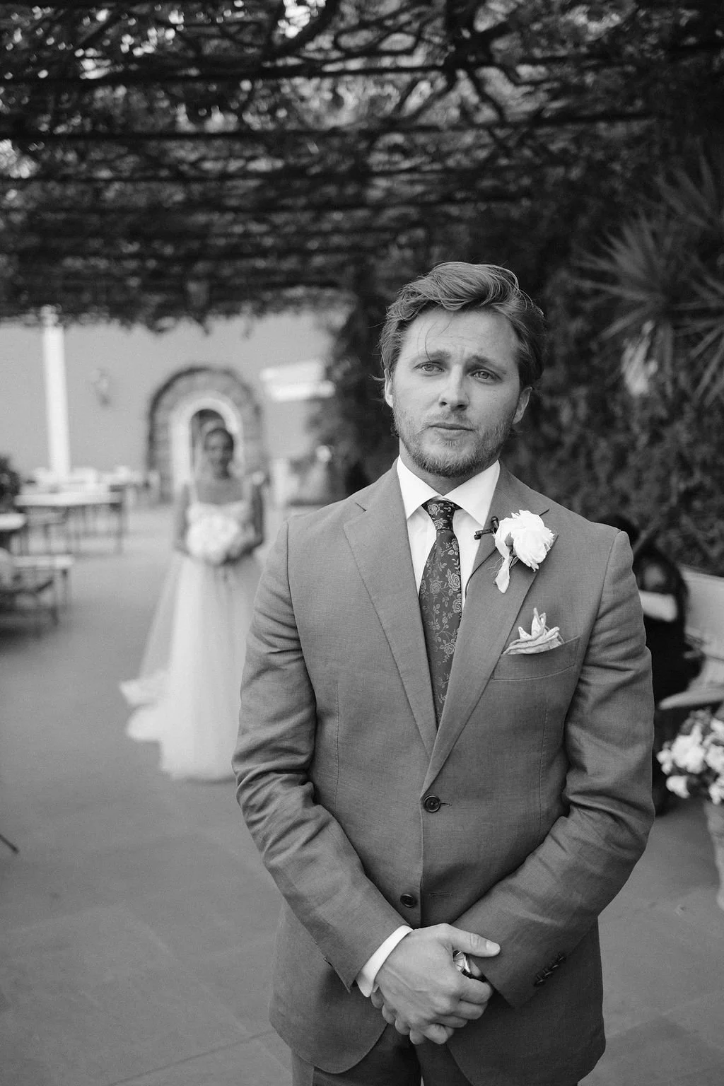 A man dressed in a suit with a floral tie and boutonniere stands outdoors with a woman in a wedding dress in the background.