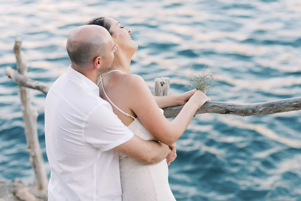 A couple standing on a wooden dock by the water, with the woman leaning back with her eyes closed and the man holding her from behind.