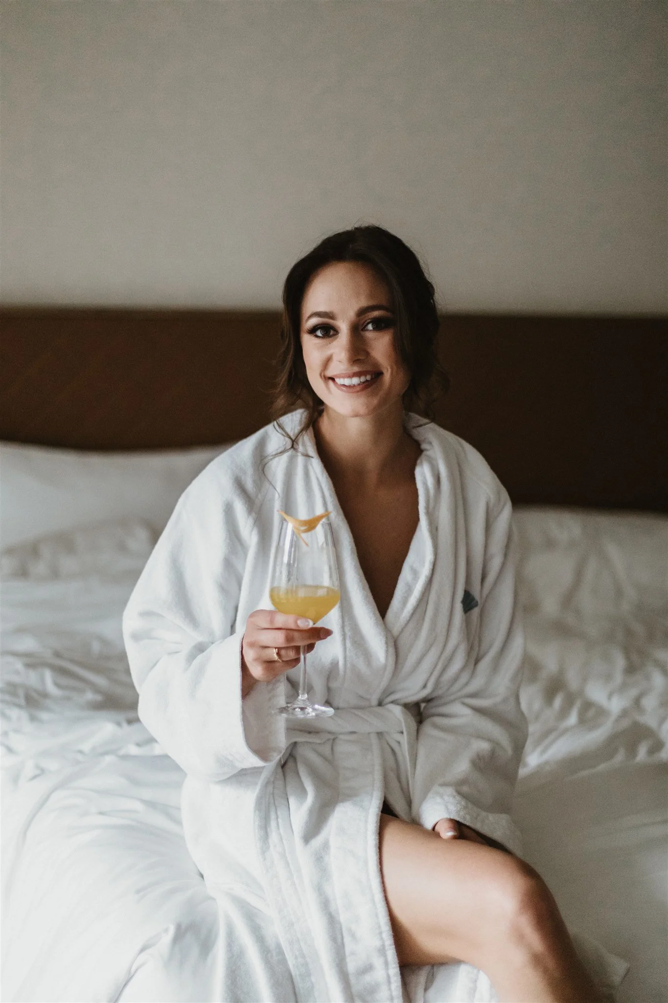 A woman sitting on a bed, wearing a white bathrobe, holding a glass of orange juice, smiling.