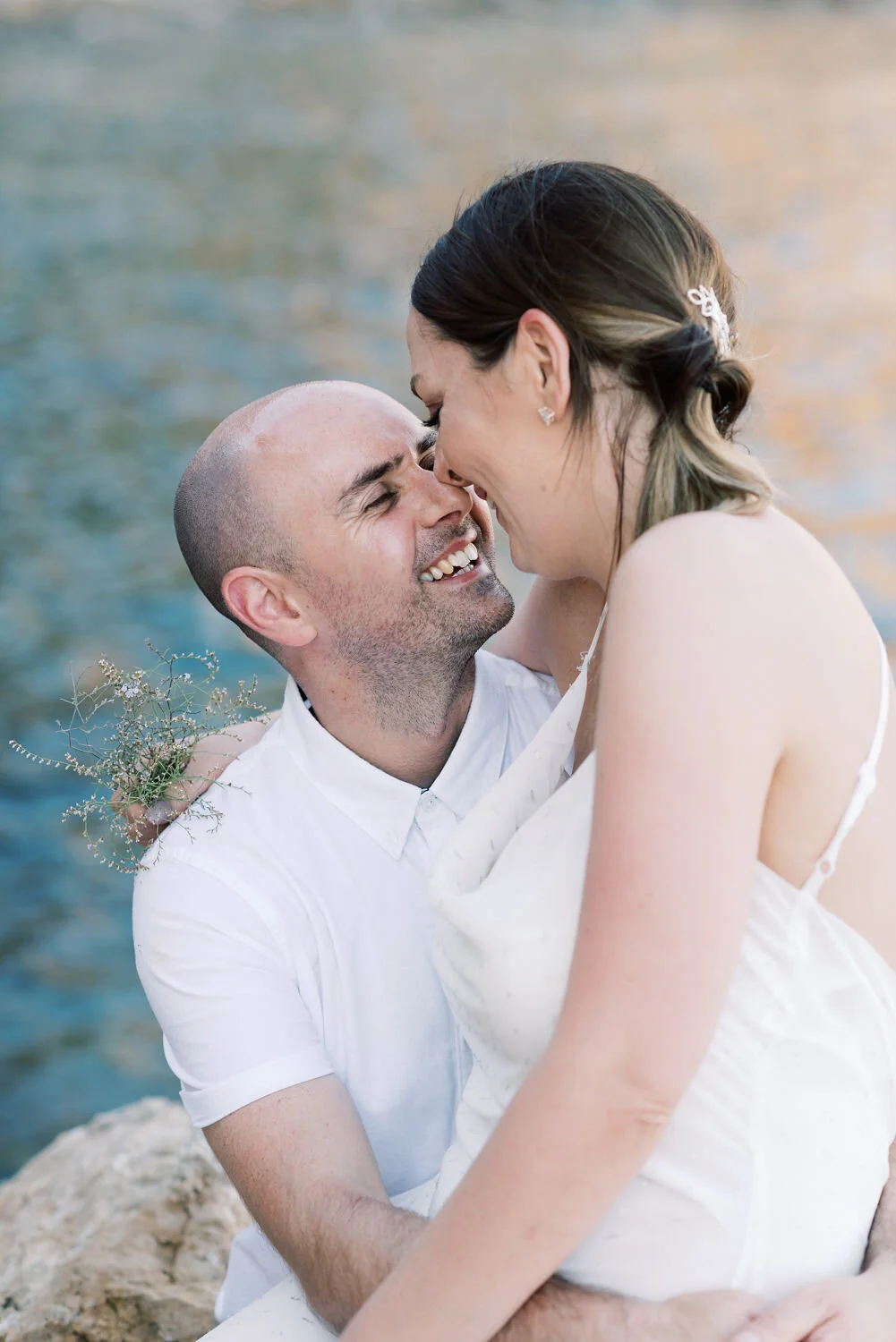 A couple celebrating a wedding or engagement near a body of water, with the man holding the woman in his arms, both smiling and their foreheads touching.