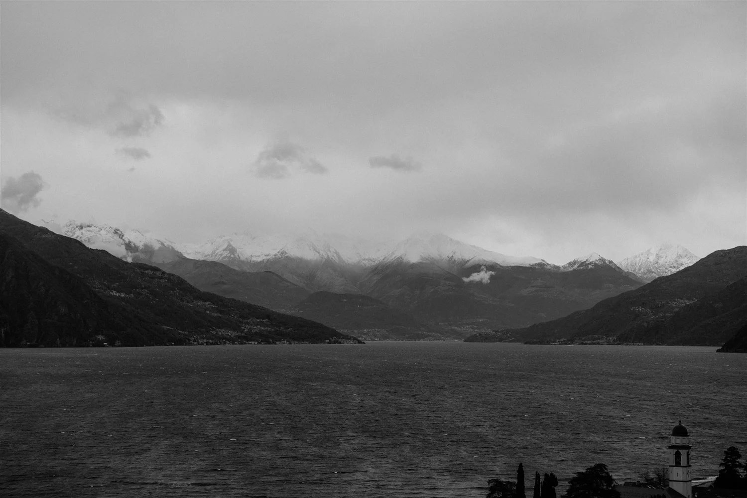 A black-and-white photograph of a mountain landscape with snow-capped peaks in the distance, a large lake in the foreground, and a lighthouse on the shoreline to the right.